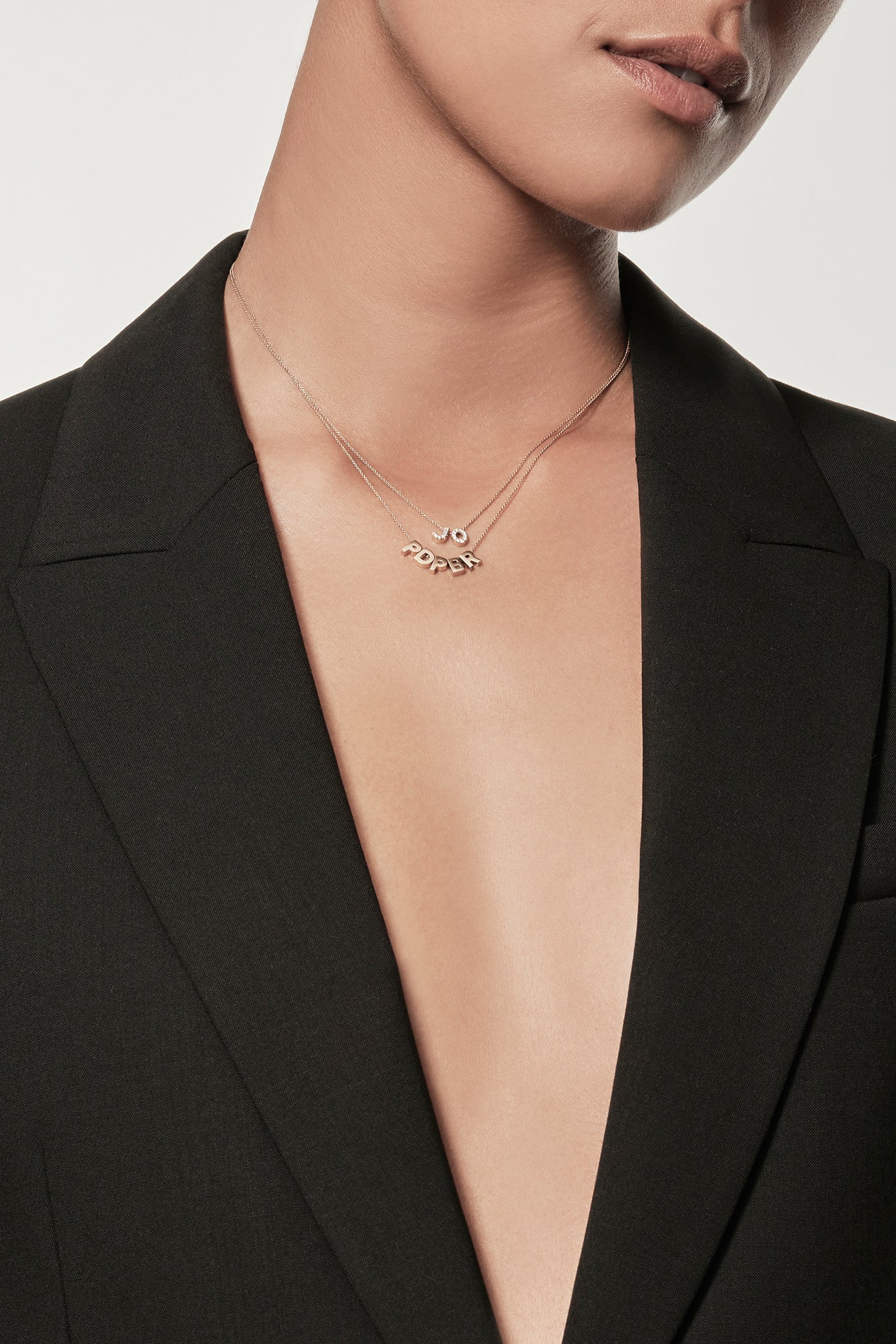 Close-up of a woman wearing a minimalist 18k gold initials “P" "D" "P" "B" "R" necklace and diamond initials "J" "O" charm necklace on a fine chain, styled with a black blazer – personalised alphabet pendant jewellery